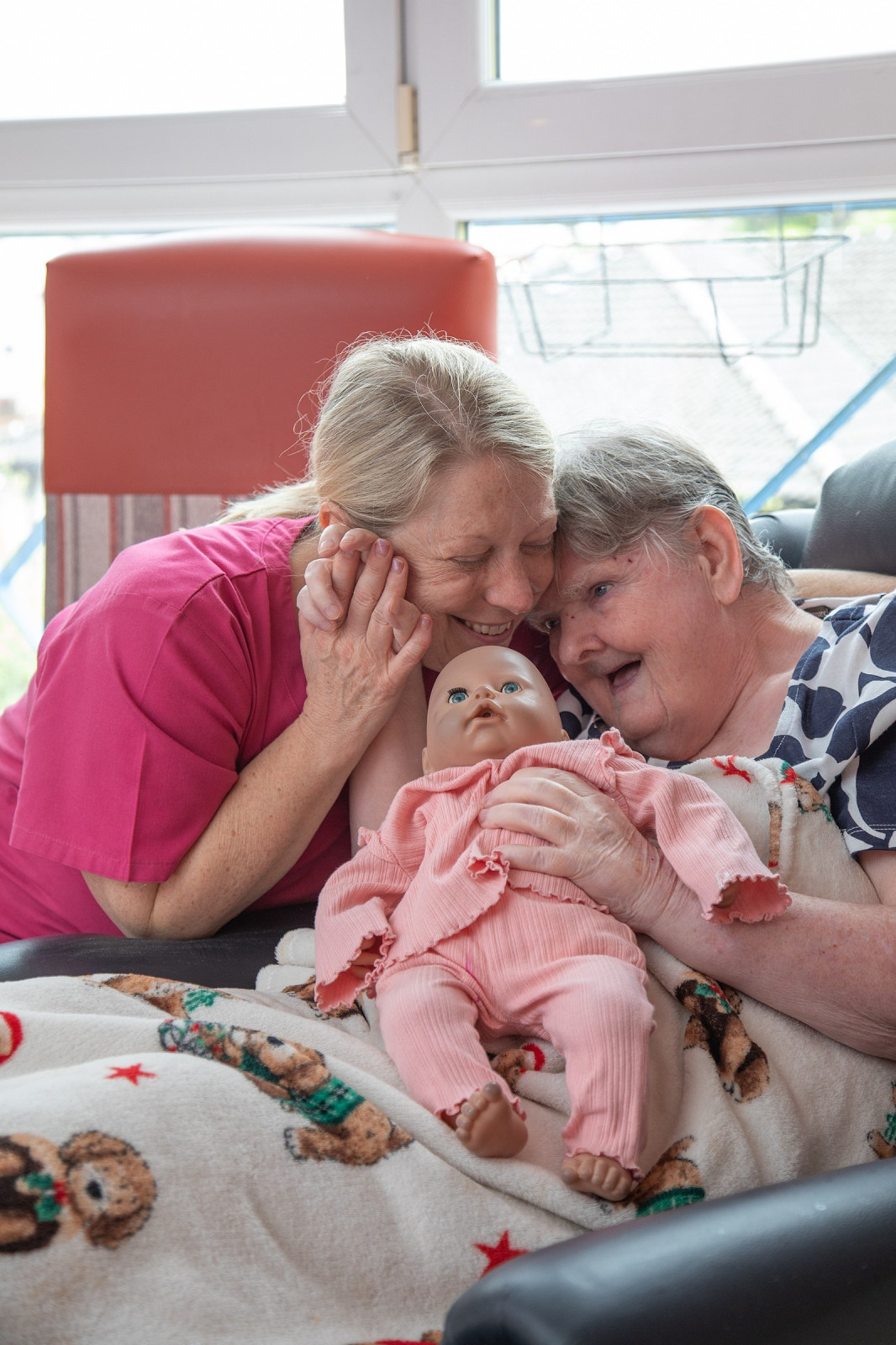A lady is in bed holding a doll. A second lady is a carer and she is leaning in to reassure the lady in the bed. Both are smiling.