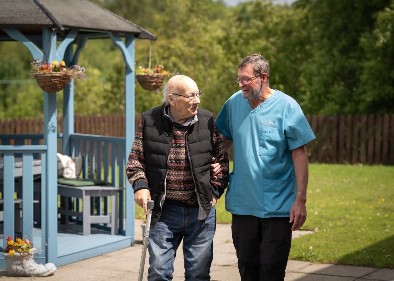 2 men walking and chatting in a garden. one has a walking stick and the other is walking arm in arm.