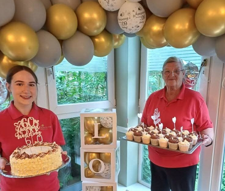 2 female carers are stood holding trays of celebration cakes for a 60th anniversary