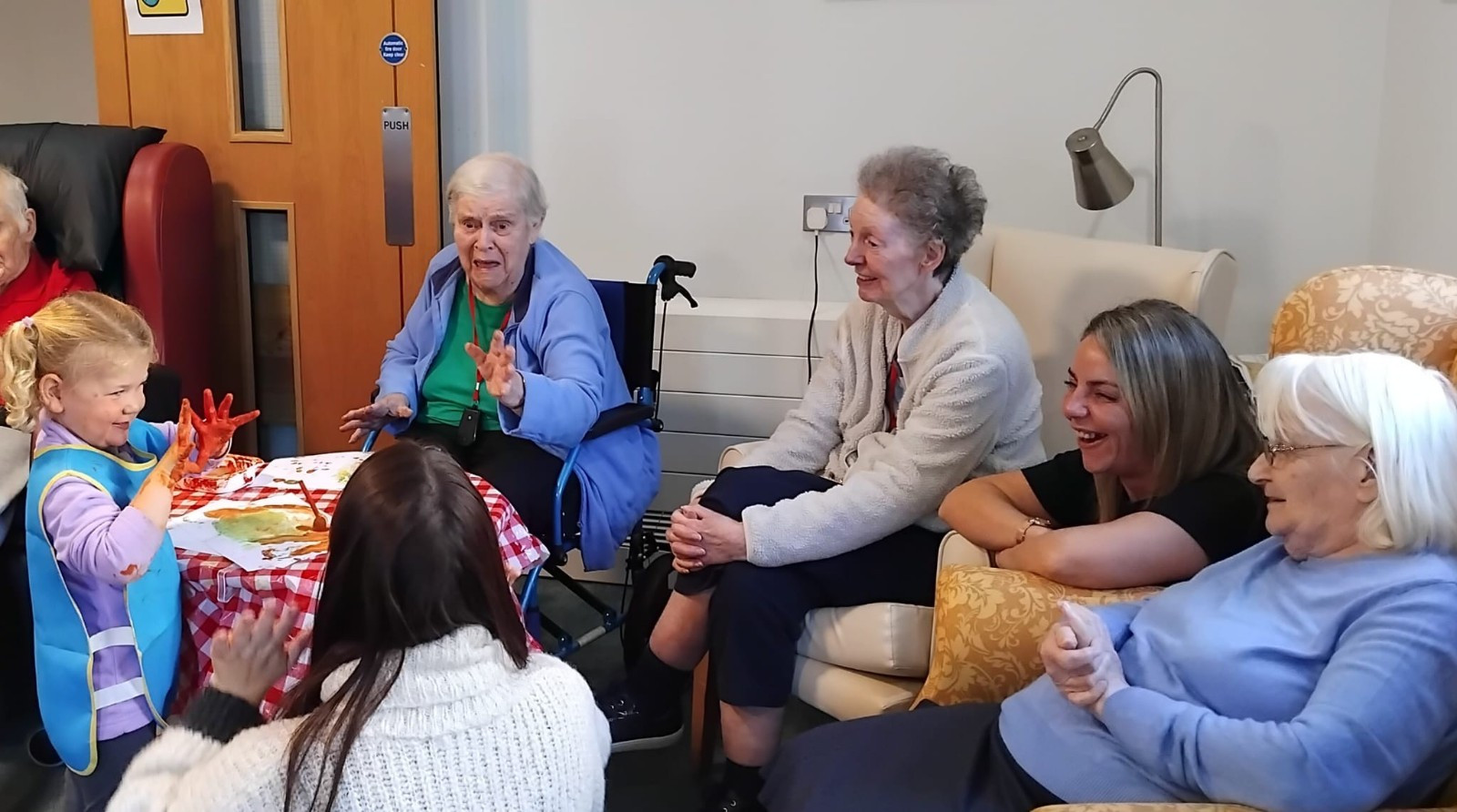 Toddler shows off her painted hands to the residents sat in a care home. All are engaged and smiling.